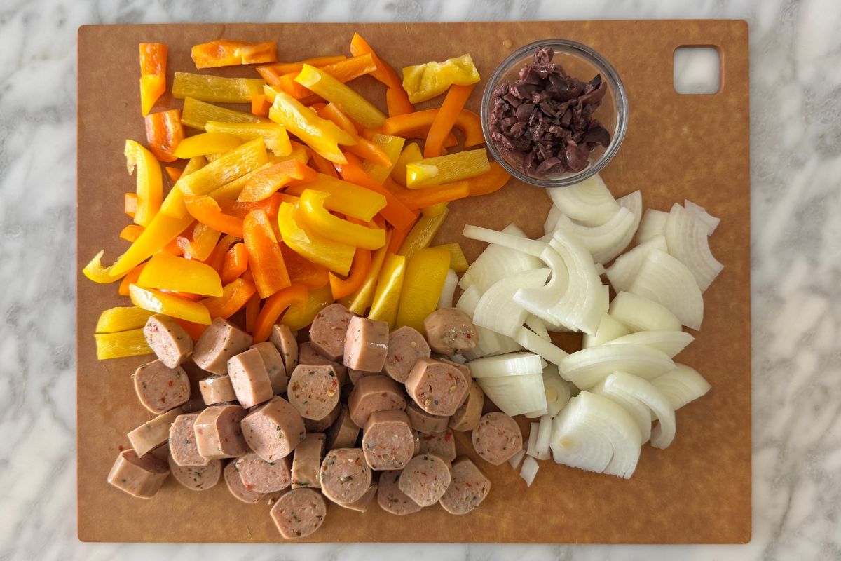 Sliced cooked sausage onions peppers and olives on a cutting board.