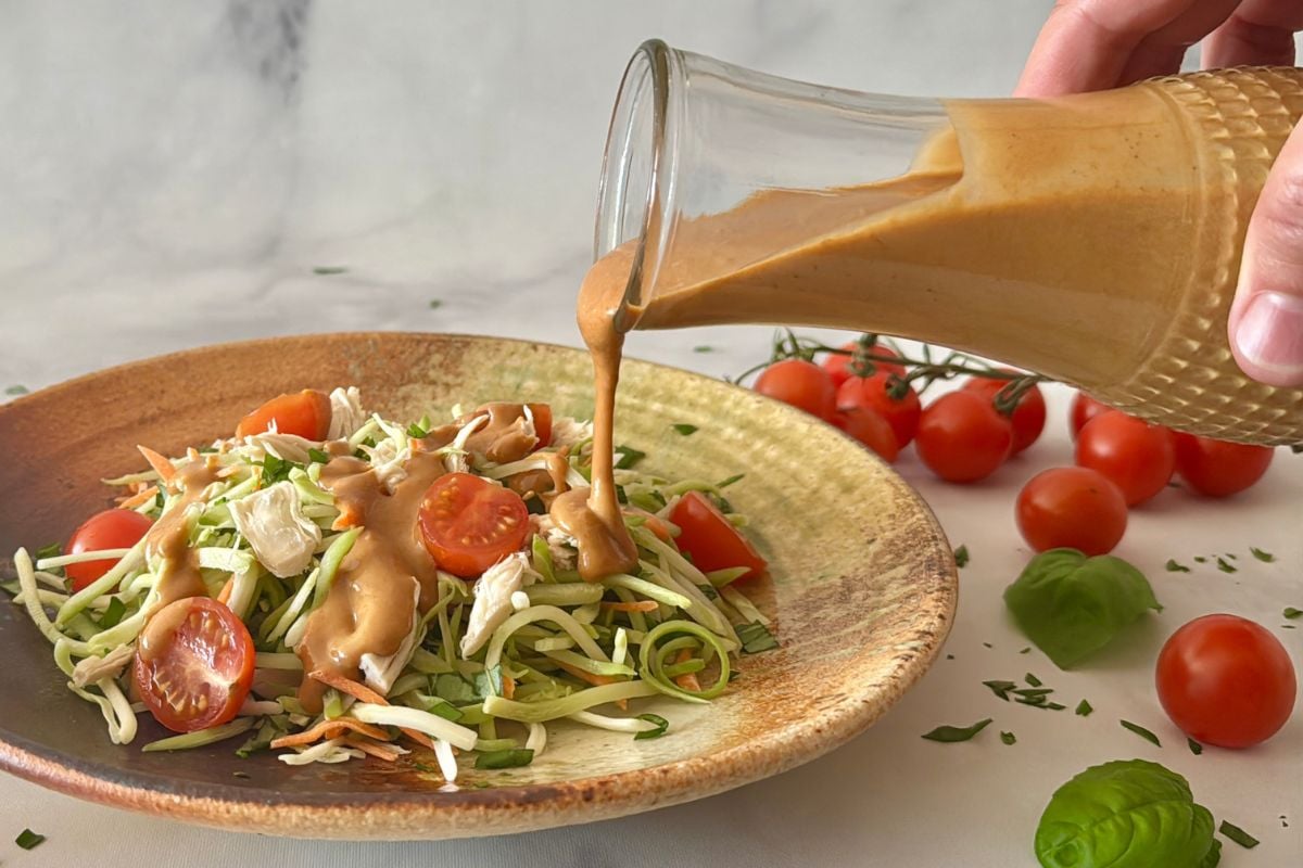 Healthy peanut dressing drizzled over a salad | Healthy Foodie Girl Creamy dressing being poured over a vegetable salad with cherry tomatoes on a ceramic plate.