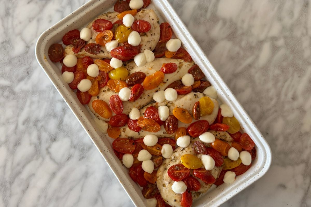 Baked chicken with tomatoes and mozzarella pearls on a sheet pan | Healthy Foodie Girl Baking dish with chicken breasts topped with roasted cherry tomatoes and mozzarella pearls on a marble surface.