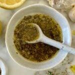 White ceramic bowl with chopped herbs in oil and a white spoon resting inside, surrounded by lemon and garlic cloves on a marble surface.