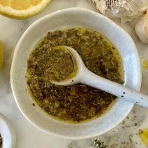 White ceramic bowl with chopped herbs in oil and a white spoon resting inside, surrounded by lemon and garlic cloves on a marble surface.