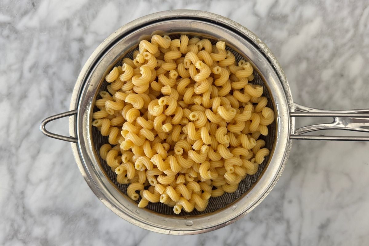 Top-down view of a stainless steel strainer filled with cooked fusilli pasta on a marble countertop.