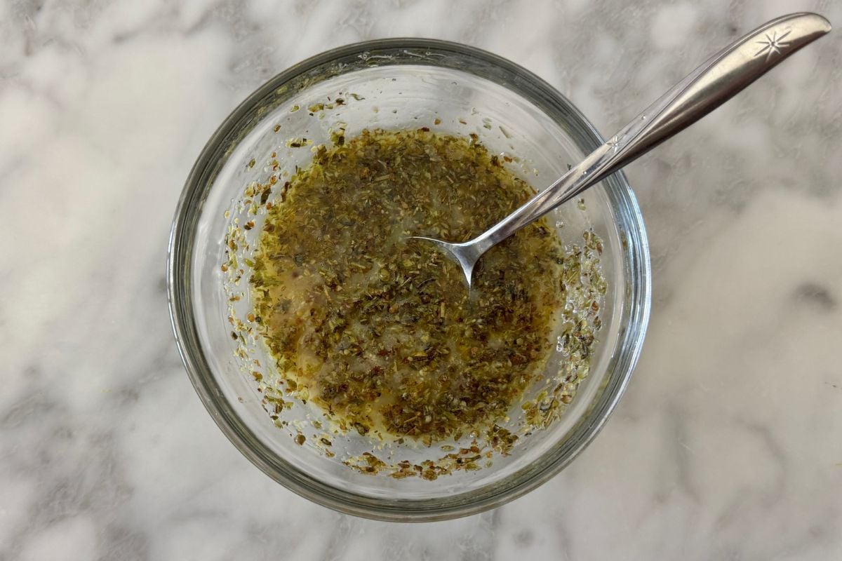 Top-down view of a glass bowl with a chopped herb mixture and a metal spoon resting inside the bowl on a marble surface