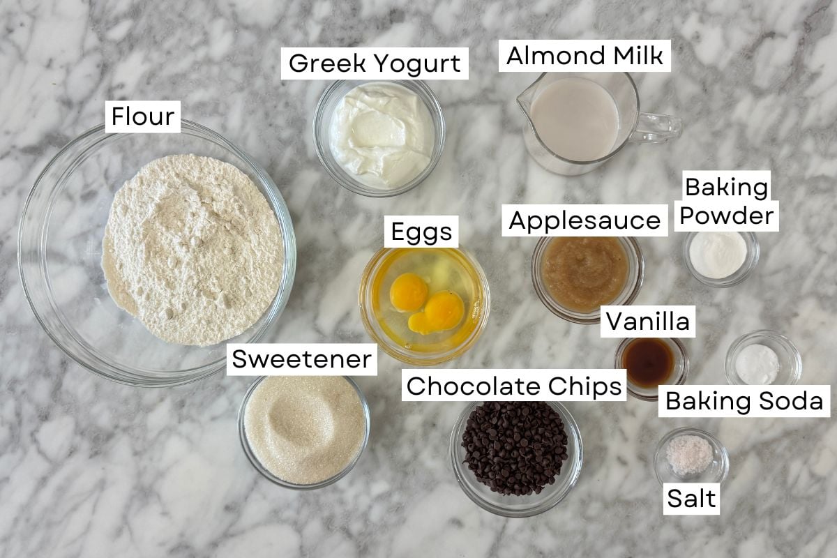 Ingredients for chocolate chip muffins with Greek yogurt in bowls | Healthy Foodie Girl Assorted baking ingredients arranged on a marble surface with labeled bowls: Flour, Greek Yogurt, Almond Milk, Eggs, Applesauce, Baking Powder, Vanilla, Baking Soda, Salt, Chocolate Chips, and Sweetener.