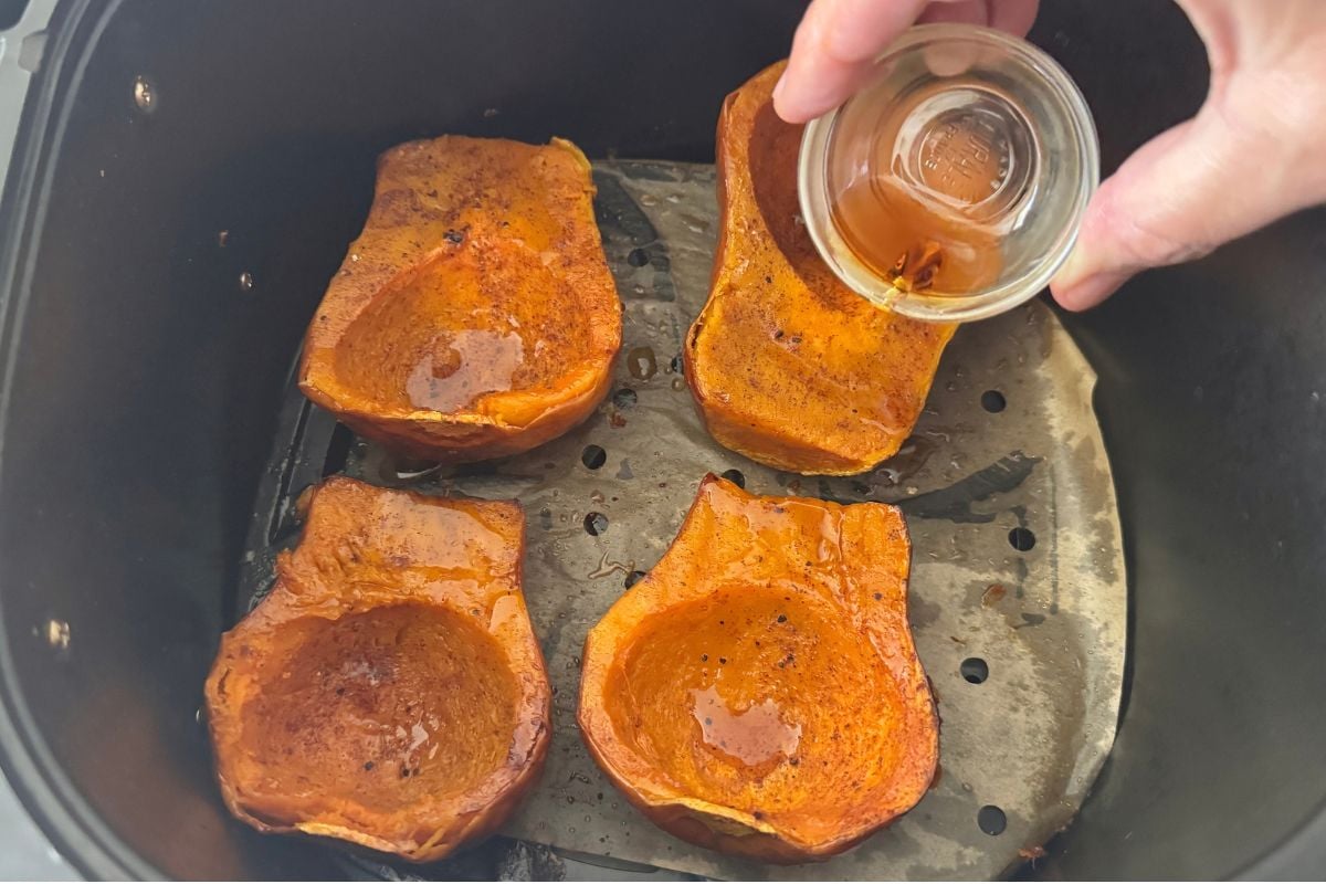 Four roasted orange squash halves arranged in an air fryer basket, seasoned and glossy with oil as a hand pours a liquid into one half.
