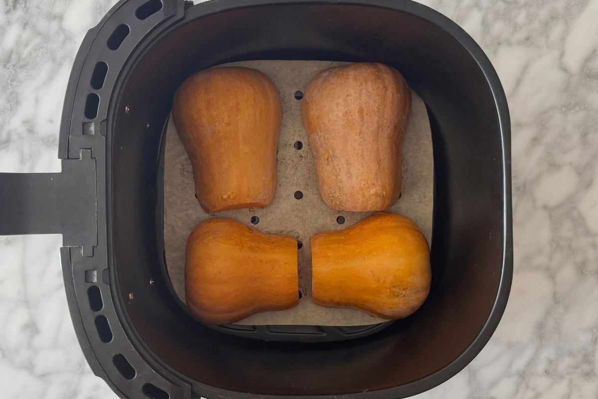 Top-down view of a black air fryer basket with four small butternut squash halves arranged in a 2x2 pattern on parchment-lined tray.