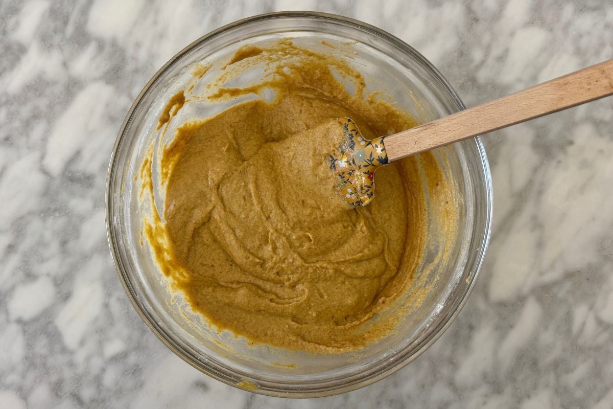 Mixed batter for sheet pan pumpkin pancakes in bowl | Healthy Foodie Girl Brown batter in a glass mixing bowl with a wooden spoon resting inside on a white marble countertop.